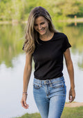 A woman with long, wavy hair is smiling and walking outdoors near a calm lake with green trees, wearing the Woolx Addie Crew Neck - Black and blue jeans.