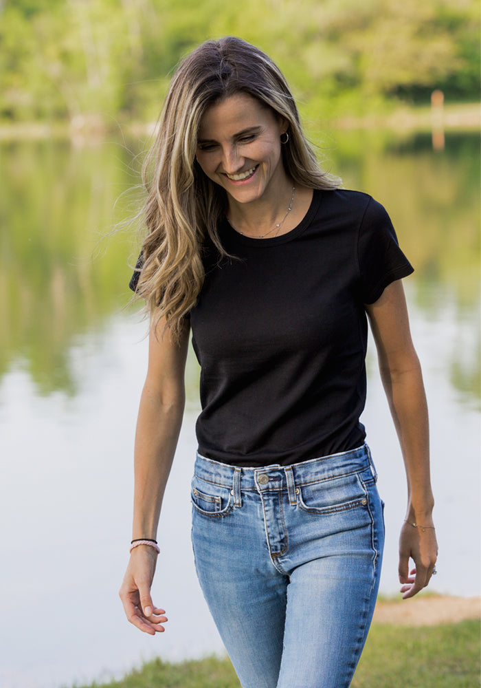 A woman with long, wavy hair is smiling and walking outdoors near a calm lake with green trees, wearing the Woolx Addie Crew Neck - Black and blue jeans.