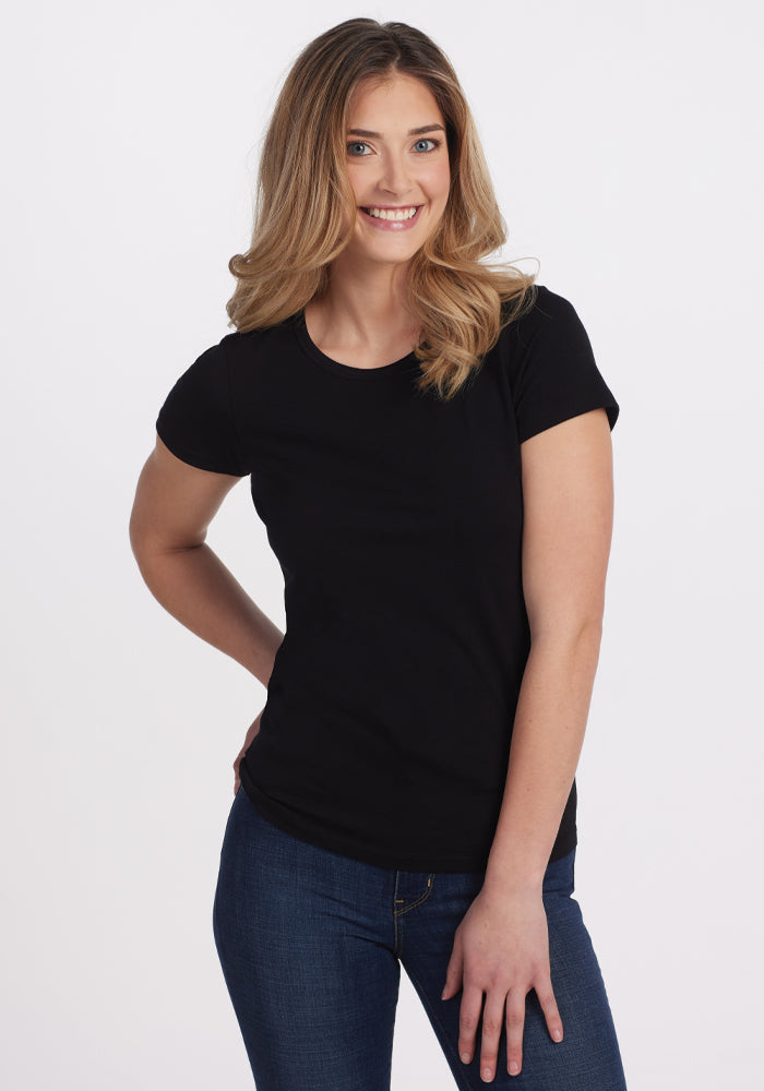 A woman with long, wavy blonde hair smiles against a white background, wearing the Woolx Addie Crew Neck in black and blue jeans, with one hand on her hip and the other by her side. | Ariana is 5'11", wearing a size S