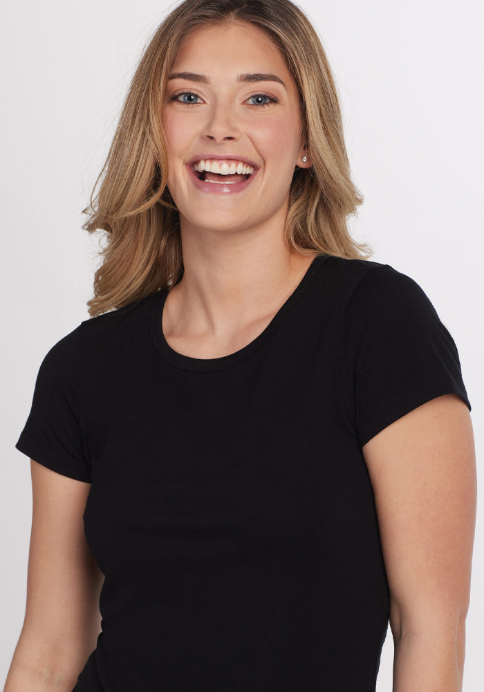 A young woman with light brown hair smiles brightly while wearing the Woolx Addie Crew Neck in black, a short-sleeved ultra-soft t-shirt, against a plain white background.