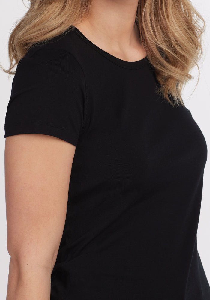 A woman with wavy blonde hair wears the Woolx Addie Crew Neck in black, a fitted, short-sleeve t-shirt made from ultra-soft, moisture-wicking fabric. She is shown from shoulders to waist, turned slightly against a plain background.