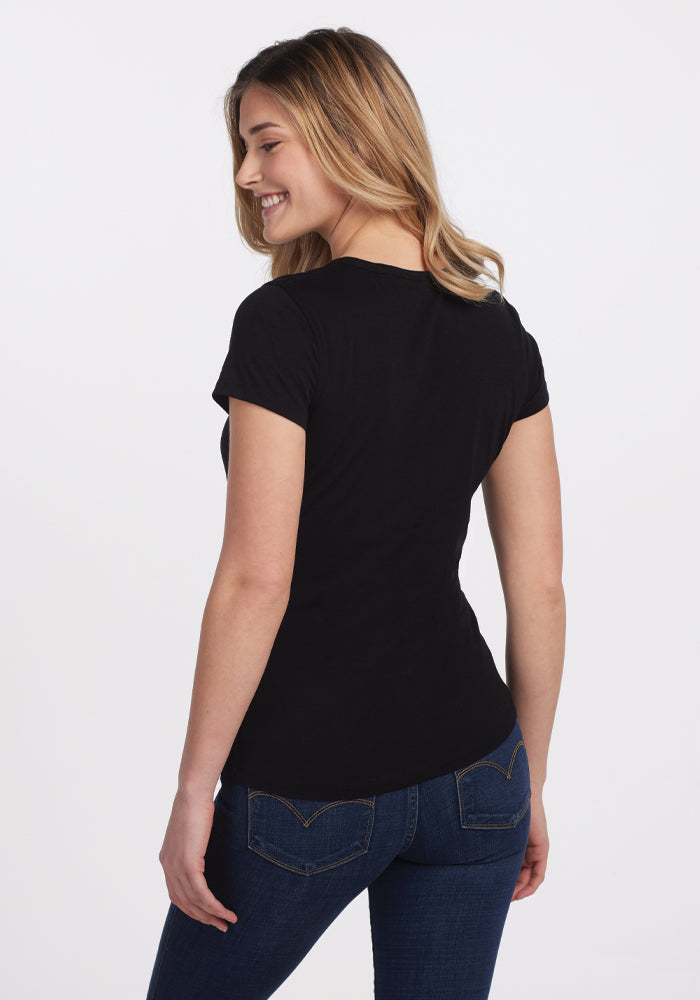 A woman with long, wavy blonde hair smiles with her back to the camera, wearing the Woolx Addie Crew Neck in black and dark blue jeans, set against a plain white background.