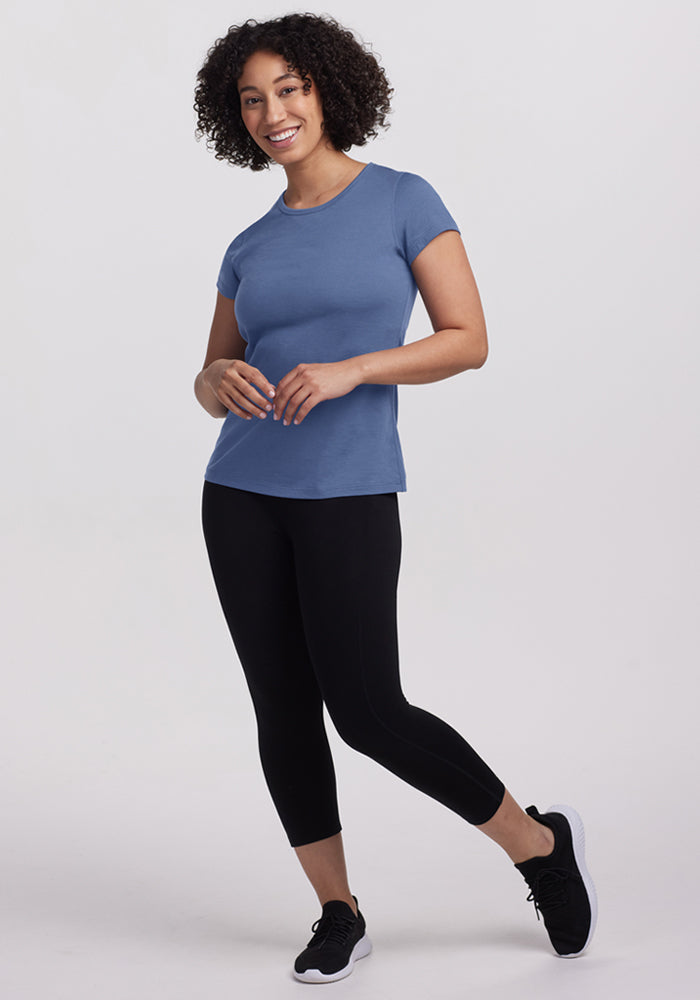 Smiling woman in a Woolx Addie Crew Neck - Skyline shirt, black leggings, and sneakers against a plain white background.