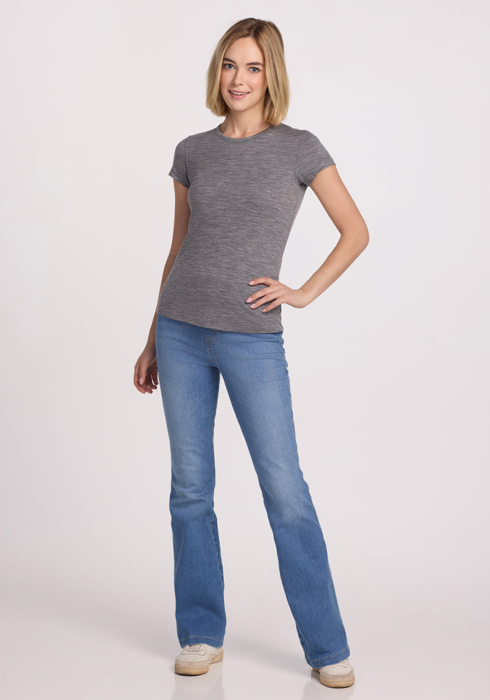 A woman with blonde hair wears the Woolx Addie Crew Neck in Graphite Heather, paired with blue jeans and white sneakers, standing with one hand on her hip and smiling against a plain white background.