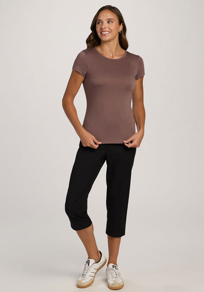 Smiling woman in a Woolx Addie Crew Neck - Peppercorn shirt, black capri pants, and white sneakers stands against a plain background.