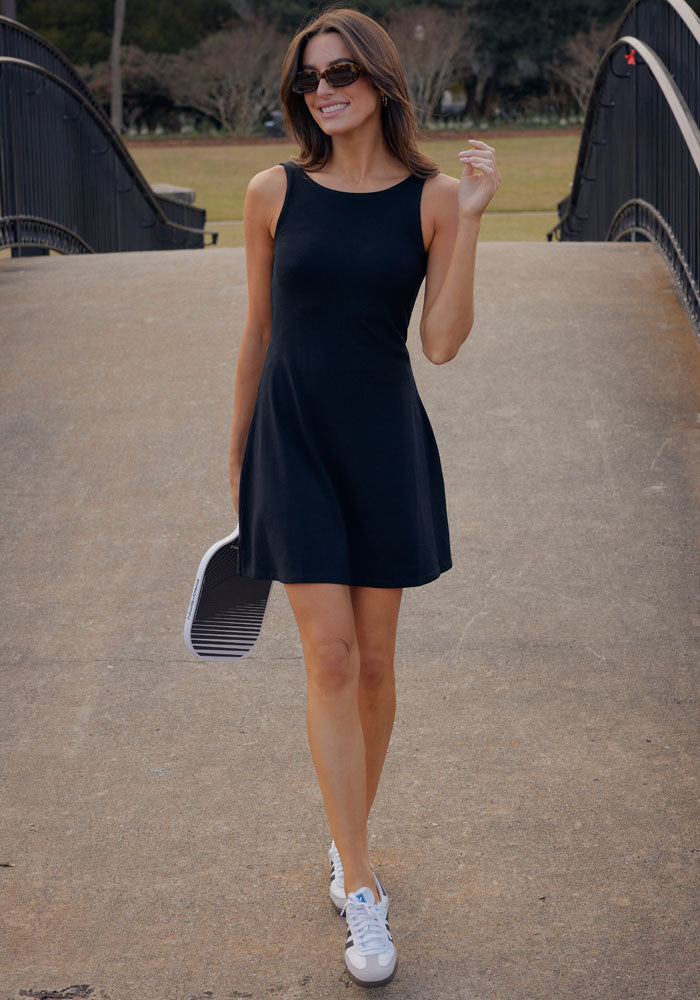 A woman in the Woolx Adrienne Active Dress - Black with built-in shorts, white sneakers, and sunglasses stands on a bridge smiling, holding a striped tote bag. She poses with one hand raised; trees and grass are in the background.