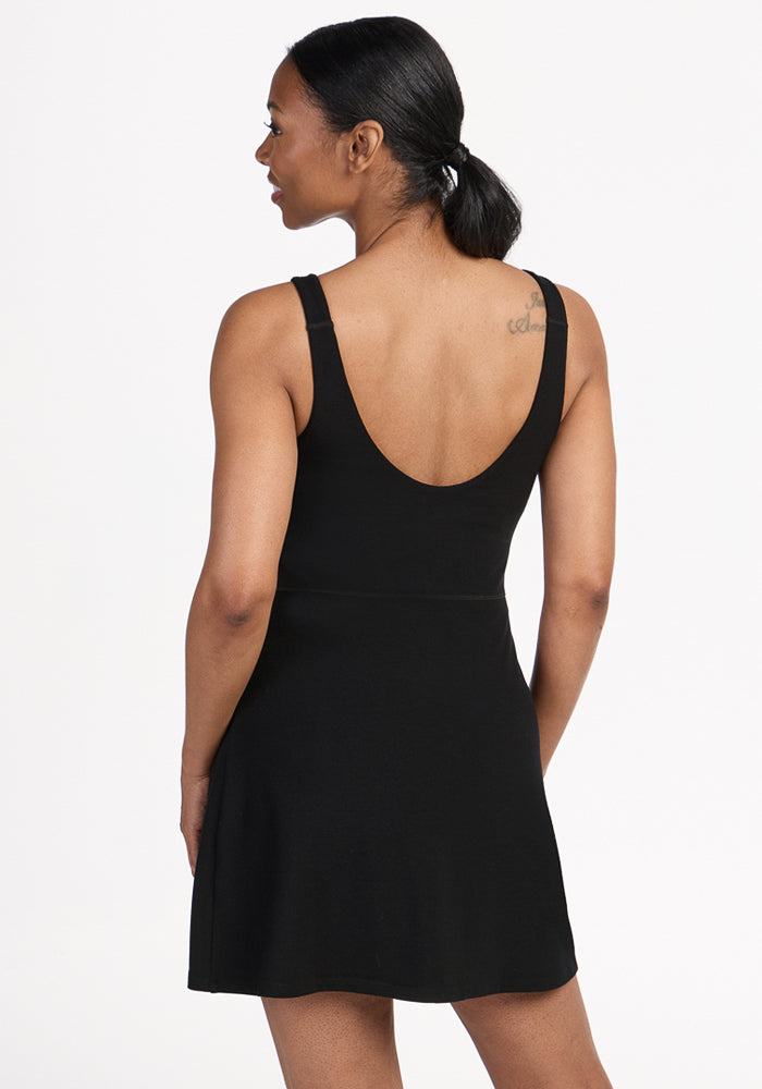 A woman with dark hair tied back wears the Woolx Adrienne Active Dress - Black, a sleeveless athletic dress with built-in shorts and a scoop back. She stands slightly sideways, revealing a small upper back tattoo against a plain white background.