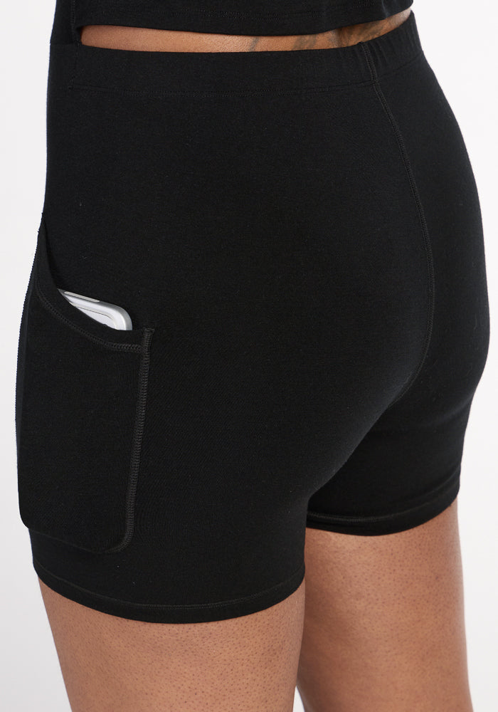 A person in black fitted shorts with a side pocket holds a smartphone, similar to how the Woolx Adrienne Active Dress - Black features built-in shorts with a pocket for easy phone storage.