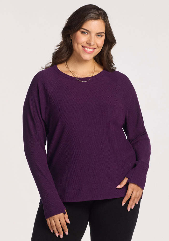 A smiling woman with long brown hair stands against a plain light background, wearing the Woolx Ainsley Sweater in Majestic Purple and black pants. She accessorizes with layered gold necklaces and has one hand on her hip.