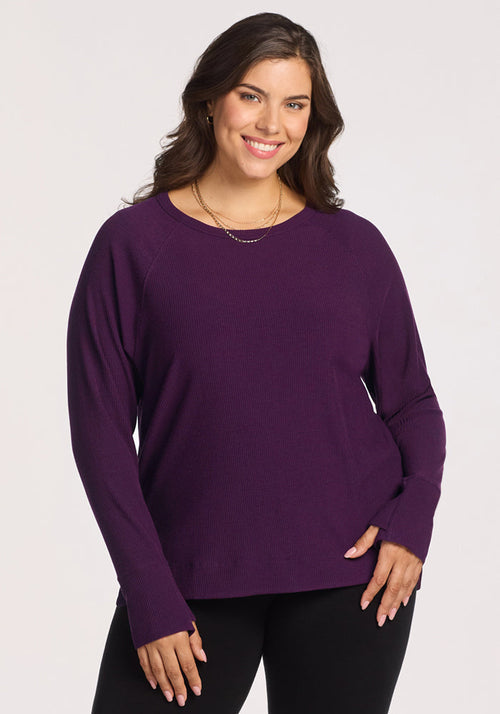 A smiling woman with long brown hair stands against a plain light background, wearing the Woolx Ainsley Sweater in Majestic Purple and black pants. She accessorizes with layered gold necklaces and has one hand on her hip.