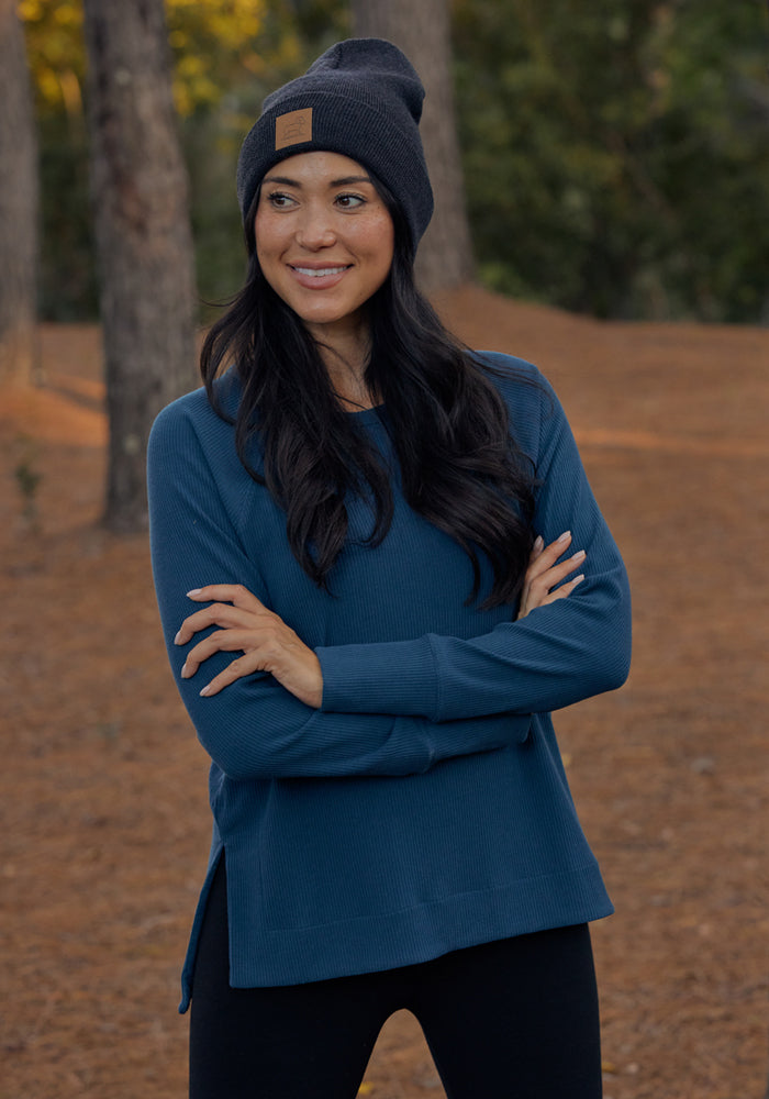 A woman with long dark hair, in a black beanie and pants, stands outdoors smiling with arms crossed, wearing the Woolx Ainsley Sweater • Final Sale - Majolica Blue. Trees and brown pine needles cover the ground behind her.