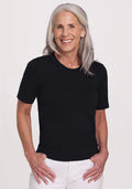 Smiling older woman with long gray hair wears the Woolx Anna Crew Sweater in black and white pants, standing against a plain light background. | Tam is 5’8.5”, wearing a size S