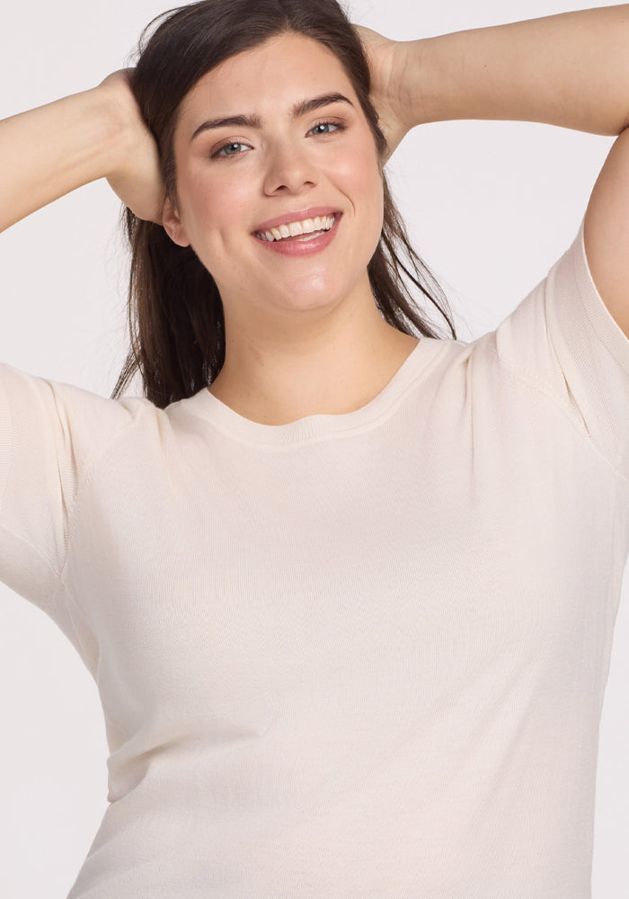 A person with long dark hair smiles confidently, wearing the Anna Crew Sweater in Dewdrop Pearl by Woolx. They casually place their hands behind their head, projecting a relaxed and cheerful vibe against a plain background.