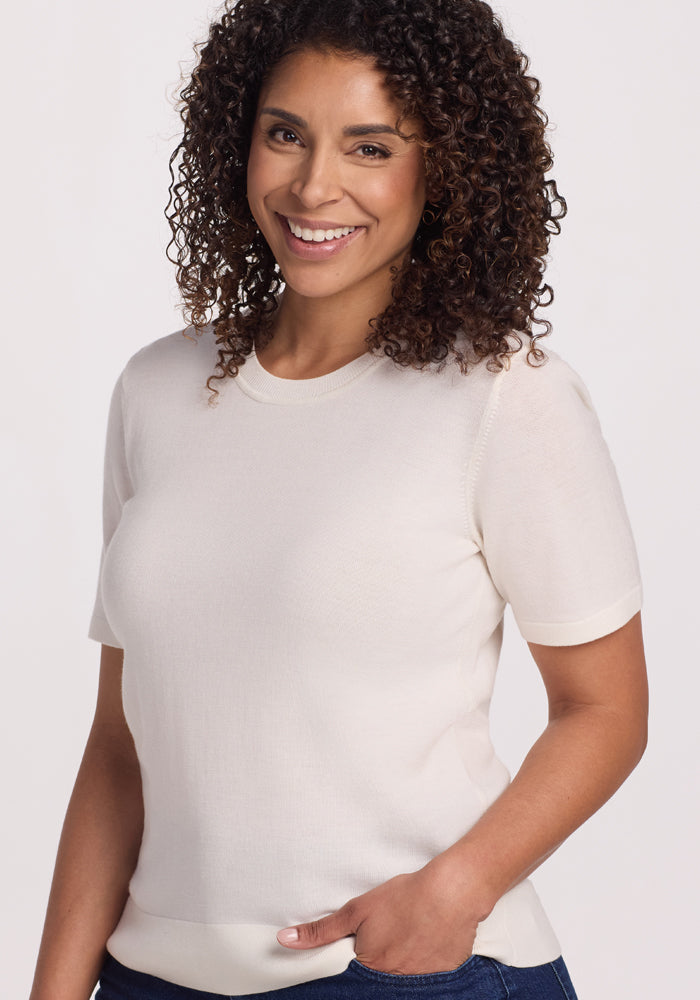 A smiling woman with curly hair wears the Anna Crew Sweater in Dewdrop Pearl by Woolx with dark jeans, standing against a plain background.