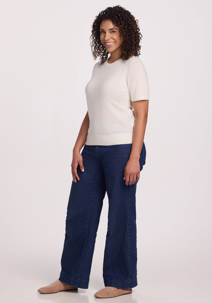 A person with curly hair smiles against a plain background, wearing a Woolx Anna Crew Sweater in Dewdrop Pearl, dark blue jeans, and beige shoes. They stand relaxed with one hand on their hip.