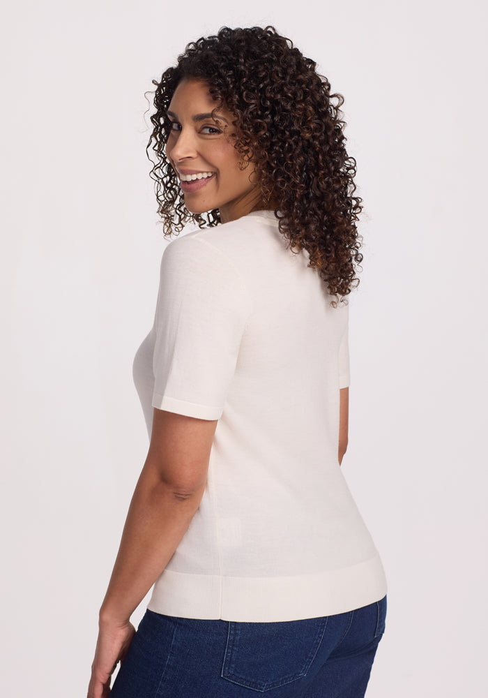 A curly-haired person smiles in profile, wearing the Woolx Anna Crew Sweater - Dewdrop Pearl and blue jeans against a plain white background.