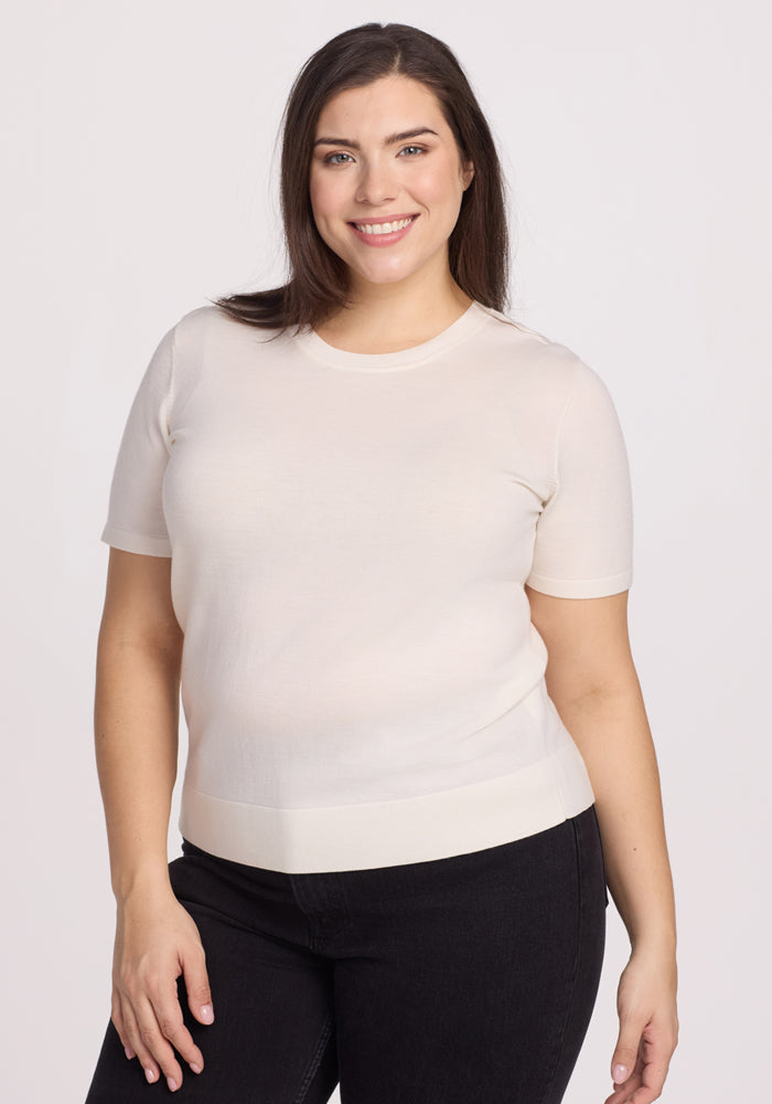 A smiling person with long dark hair wears the Woolx Anna Crew Sweater in Dewdrop Pearl, paired with dark pants, standing against a plain light background. | Juliana is 5'10", wearing a size XL