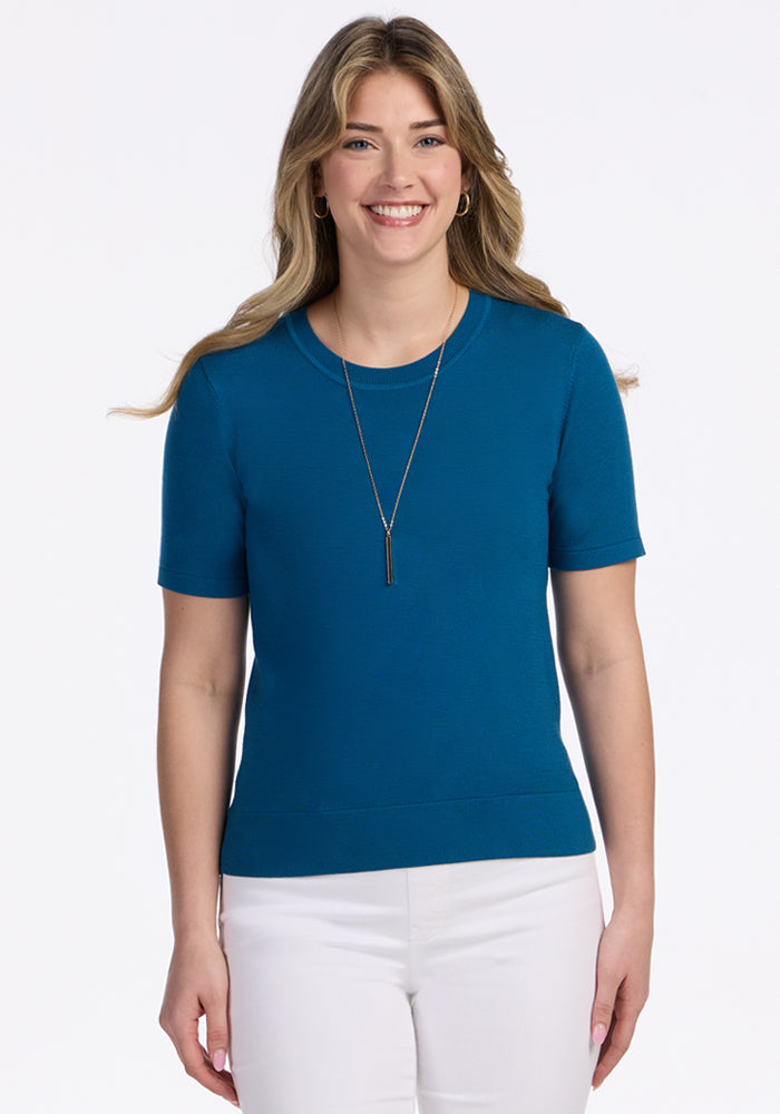 A woman with long, wavy blonde hair smiles while wearing the Woolx Anna Crew Sweater in Moroccan Blue, paired with white pants and a pendant necklace, standing against a plain white background. | Ariana is 5'11", wearing a size S