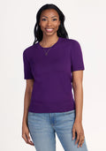 A woman with long dark hair smiles at the camera, wearing the Woolx Anna Crew Sweater - Plum Crush paired with light blue jeans and a gold necklace, standing in a relaxed pose against a plain light background. | Jade is 5’10”, wearing a size S