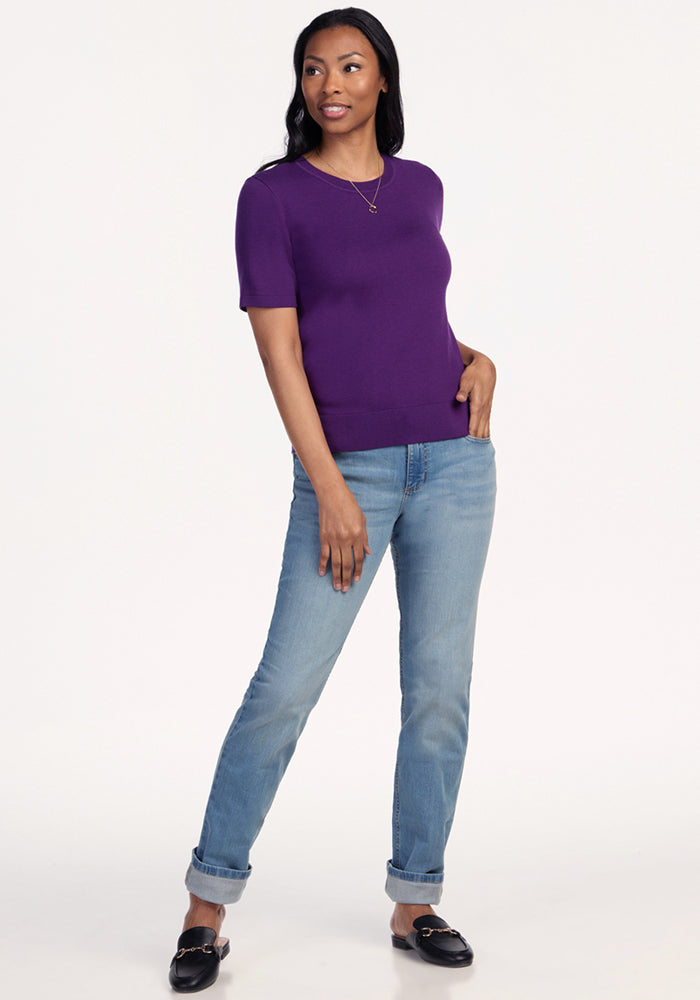 A woman smiles and looks to the side, wearing the Woolx Anna Crew Sweater in Plum Crush, light blue jeans with rolled cuffs, black loafers, and a necklace against a plain background, with one hand in her pocket.