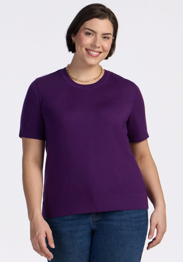 A woman with short brown hair smiles while wearing the Woolx Anna Crew Sweater in Plum Crush, paired with blue jeans and a gold chain necklace, standing against a plain white background. | Juliana is 5'10", wearing a size XL