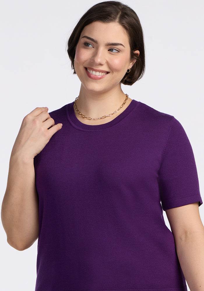 A woman with short brown hair smiles while wearing the Woolx Anna Crew Sweater in Plum Crush and a gold chain necklace against a plain light background.