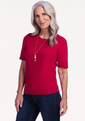 A smiling older woman with long gray hair wears the Woolx Anna Crew Sweater in Poppy Spark, dark jeans, and a long necklace, standing against a plain light background. | Tam is 5’8.5”, wearing a size S