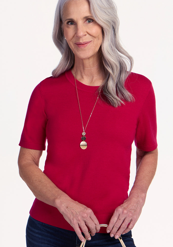 A smiling older woman with long, wavy gray hair wears the Woolx Anna Crew Sweater in Poppy Spark and dark jeans, accessorized with a pendant necklace, standing against a plain light background.