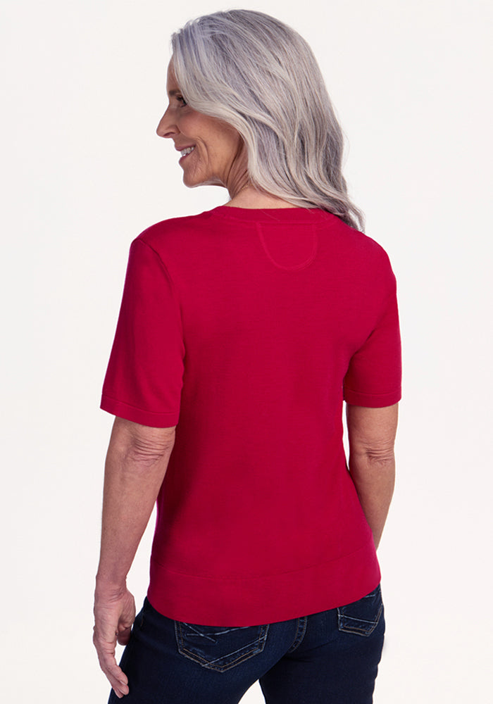 An older woman with long gray hair smiles, standing with her back to the camera in a Woolx Anna Crew Sweater – Poppy Spark and dark jeans against a light background.