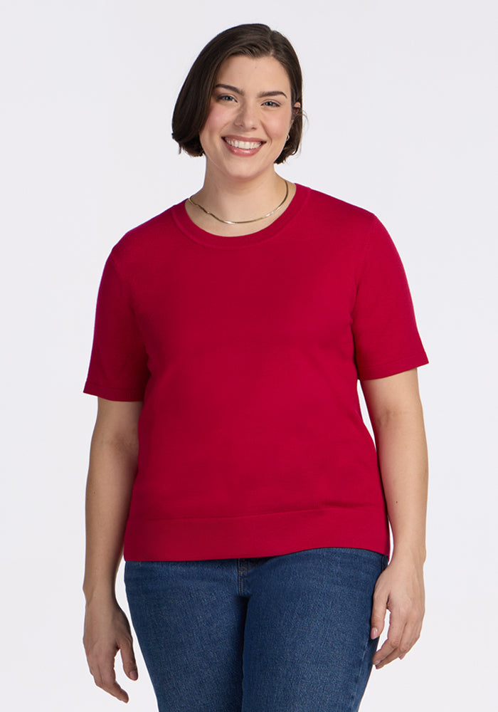 A woman with short brown hair smiles against a white background, wearing the Woolx Anna Crew Sweater in Poppy Spark—a chic short-sleeve style—paired with classic blue jeans. | Juliana is 5'10", wearing a size XL