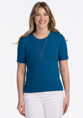 A woman with long, wavy blonde hair smiles while wearing the Woolx Anna Crew Sweater in Moroccan Blue, paired with white pants and a pendant necklace, standing against a plain white background. | Ariana is 5'11", wearing a size S