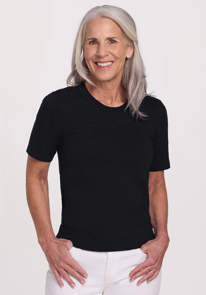 Smiling older woman with long gray hair wears the Woolx Anna Crew Sweater in black and white pants, standing against a plain light background. | Tam is 5’8.5”, wearing a size S