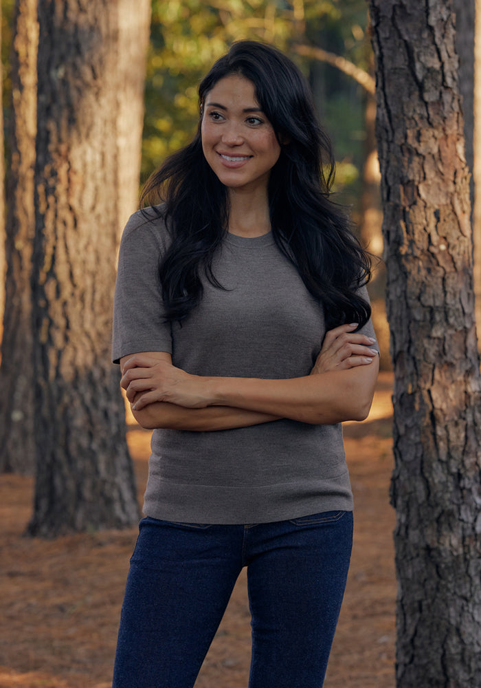 A woman with long dark hair stands outdoors among tall trees, smiling and looking to the side. She is wearing the Woolx Anna Crew Sweater in Simply Taupe and blue jeans, with her arms crossed. Pine needles cover the ground.