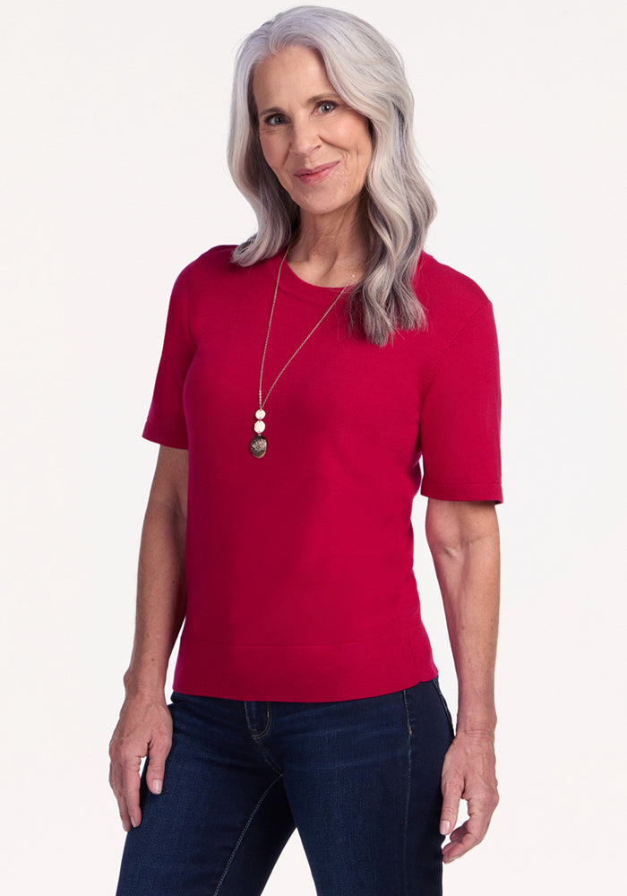 A smiling older woman with long gray hair wears the Woolx Anna Crew Sweater in Poppy Spark, dark jeans, and a long necklace, standing against a plain light background. | Tam is 5’8.5”, wearing a size S