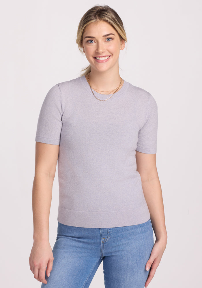 A blonde woman with her hair tied back smiles against a light background, wearing the Woolx Anna Crew Sweater in Mist (Final Sale), blue jeans, and a layered gold necklace.