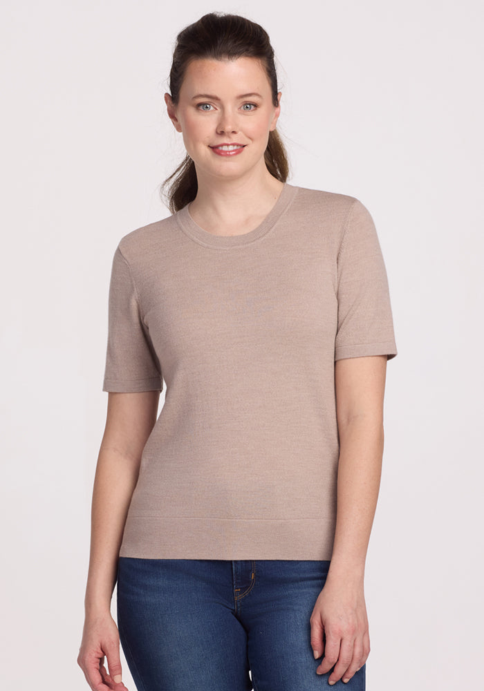 A woman with long brown hair tied back wears the Woolx Anna Crew Sweater in Sandstone Haze with blue jeans, standing and smiling at the camera against a plain light background.