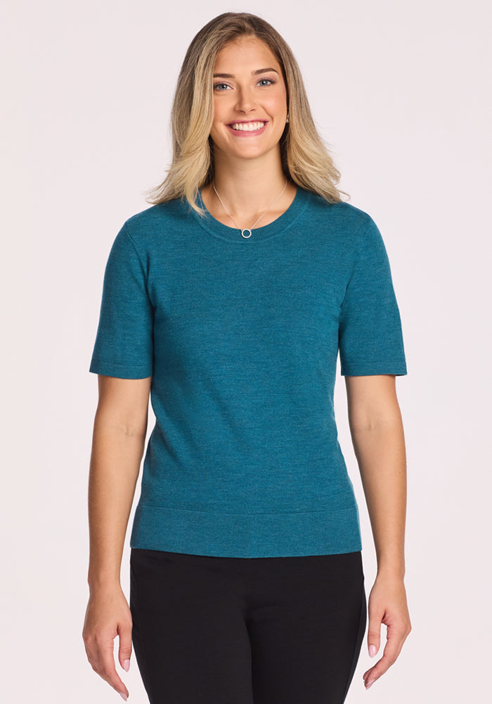 A woman with shoulder-length blonde hair, wearing the Woolx Anna Crew Sweater in Harbor Teal and black pants, smiles against a plain light background.