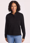 A woman with curly hair smiles at the camera, wearing the Woolx Annie 3 Button Pullover in black with light blue jeans, standing against a plain light background. | Tori is 5'7", wearing a size S