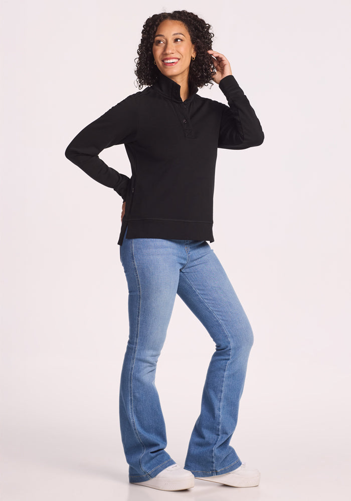 A woman with curly hair smiles while posing in the Woolx Annie 3 Button Pullover in black, blue flared jeans, and white sneakers against a plain white background.