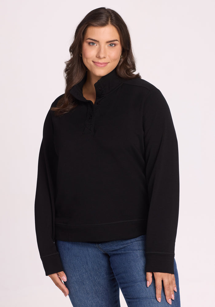 A woman with long brown hair, wearing the Woolx Annie 3 Button Pullover in black and blue jeans, stands against a plain light background and smiles softly with one hand by her side. | Juliana is 5'10", wearing a size XL