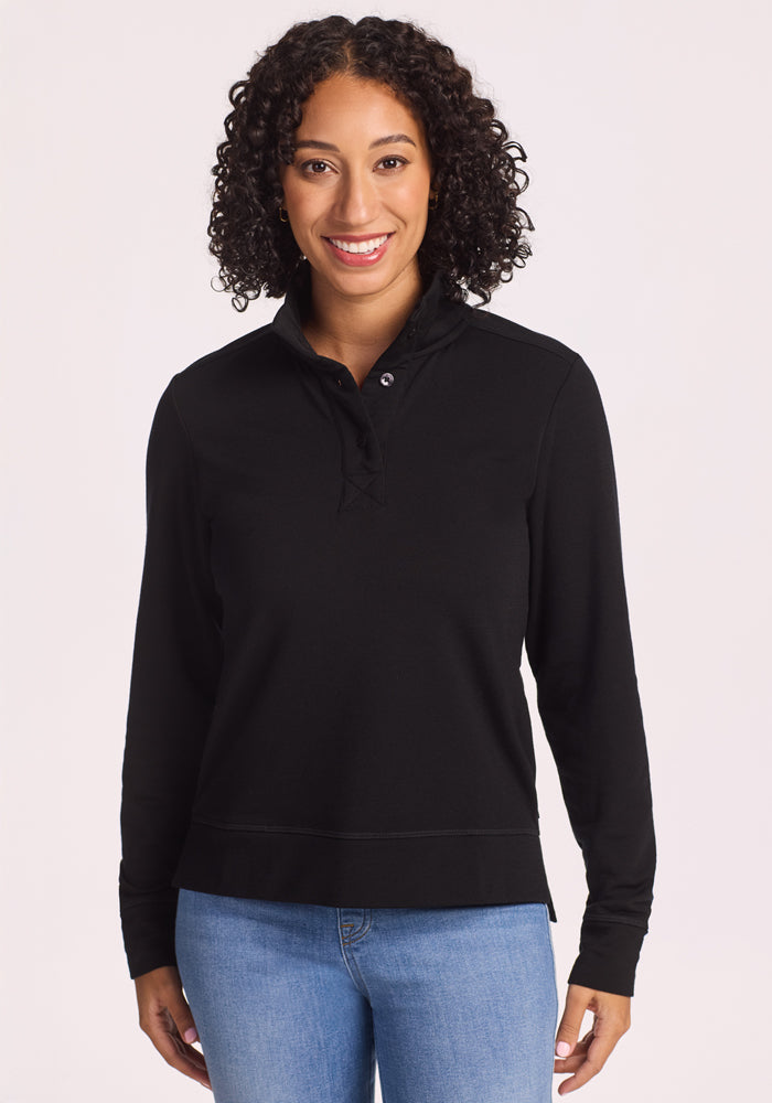A woman with curly hair smiles at the camera, wearing the Woolx Annie 3 Button Pullover in black with light blue jeans, standing against a plain light background. | Tori is 5'7", wearing a size S