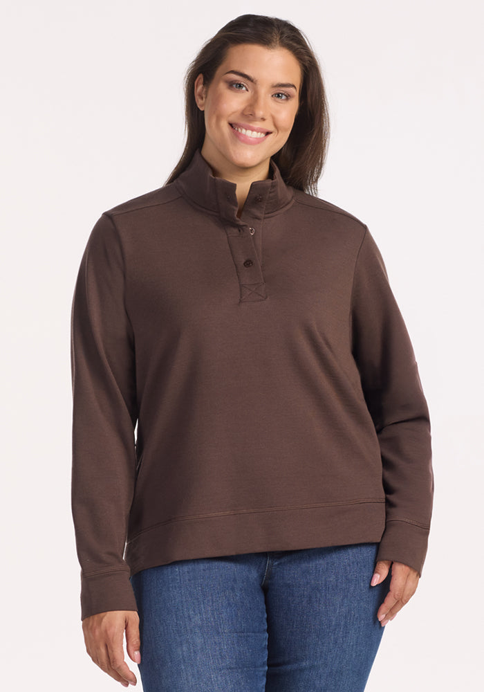 A woman with long brown hair smiles while wearing the Woolx Annie 3 Button Pullover in Bracken and blue jeans, standing against a plain light background.