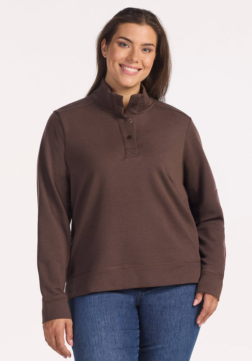 A woman with long brown hair smiles while wearing the Woolx Annie 3 Button Pullover in Bracken and blue jeans, standing against a plain light background.