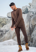A man in Woolx Arctic Leggings • Final Sale - Bourbon Oak stands smiling on snowy, rocky terrain, looking down as he adjusts his sleeve, wearing tan hiking boots.