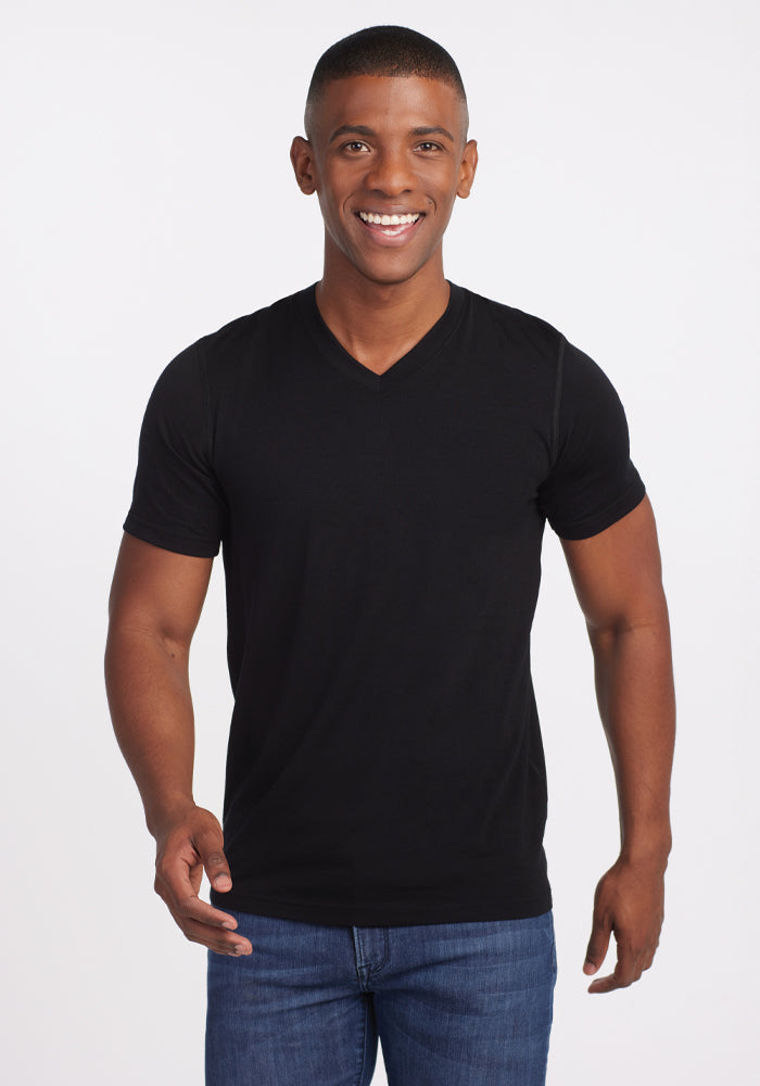 A person smiling and standing against a plain white background, wearing the Ashton V Neck in black by Woolx paired with blue jeans, enjoying the comfort of the lightweight, odor-free Merino fabric. | Trell is 6’2”, wearing a size M