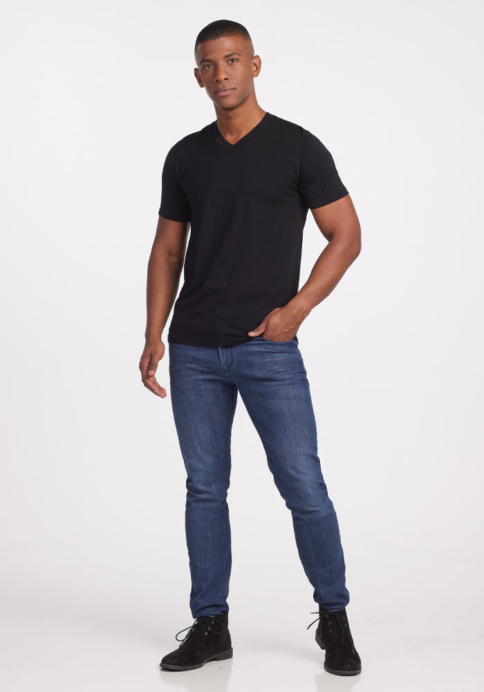 A person stands against a plain white background, wearing the Ashton V Neck in black by Woolx, paired with blue jeans and black shoes. The odor-free shirt complements their neutral expression as their hands rest relaxed by their sides.