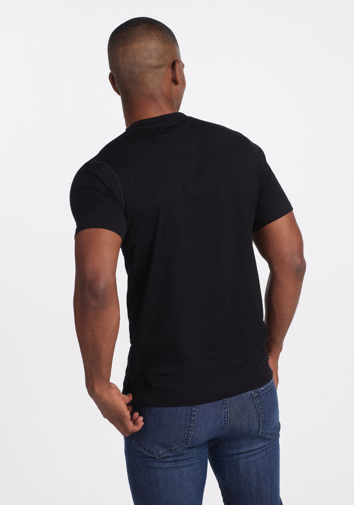 A man stands with his back to the camera, wearing the Ashton V Neck - Black by Woolx paired with blue jeans. The lightweight fabric of this T-shirt ensures comfort and is designed to be odor-free. Set against a plain white background, his relaxed posture, with one arm slightly bent, adds a casual vibe.