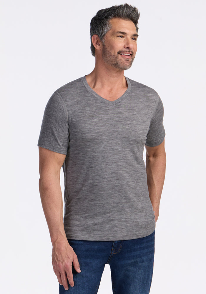 A man with short dark and gray hair and a beard is smiling, wearing the Woolx Ashton V Neck in Graphite Heather and blue jeans, standing against a plain white background. | Matthew is 6’, wearing a size L