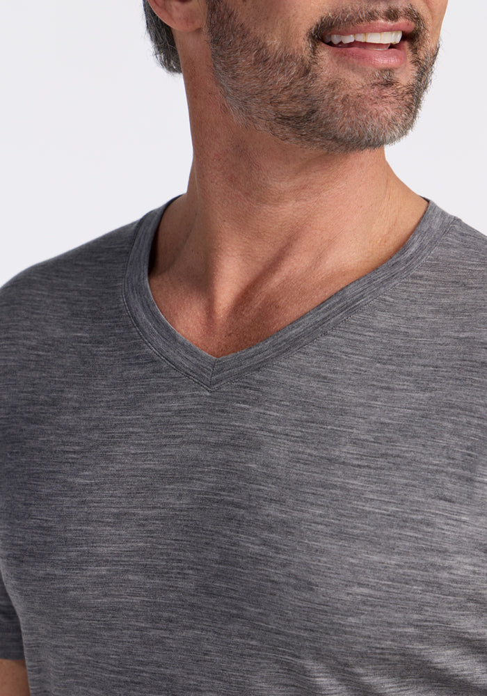 A close-up of a man wearing the Woolx Ashton V Neck in Graphite Heather. Only his lower face, neck, and upper torso are shown. He has a short beard, a slight smile, and stands against a plain light background.
