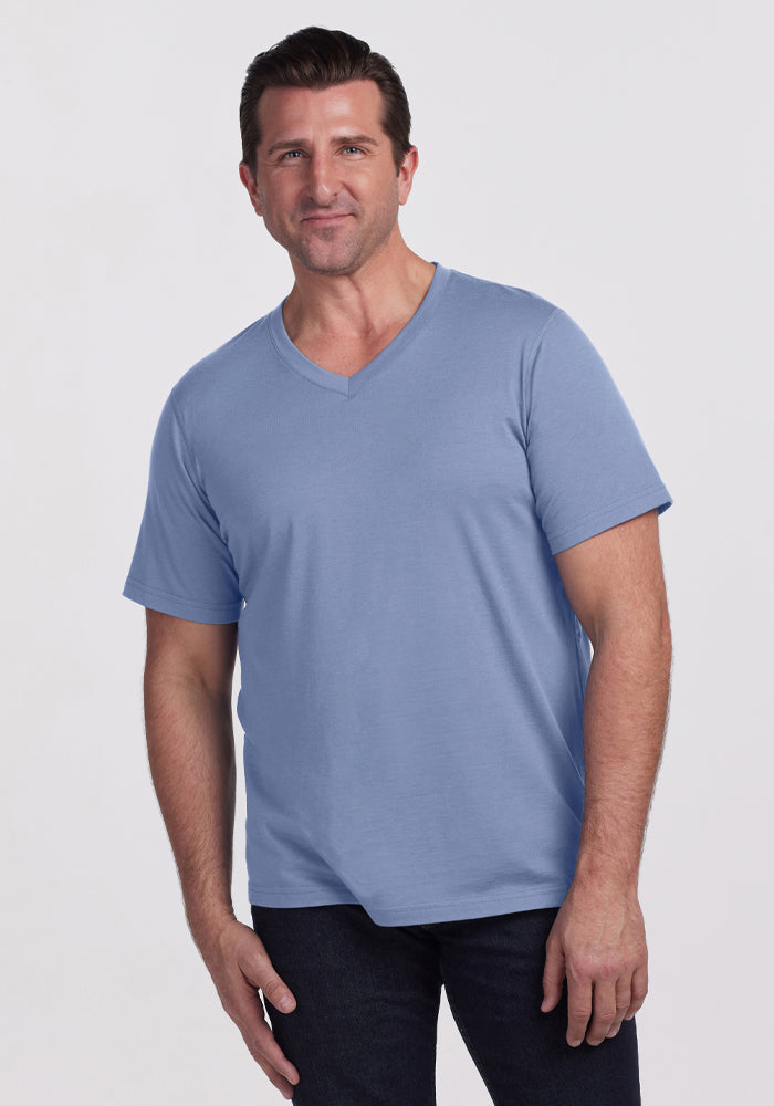 A man smiles against a white background, wearing the Woolx Ashton V Neck • Final Sale - Stonewash tee with dark pants and one hand relaxed at his side.